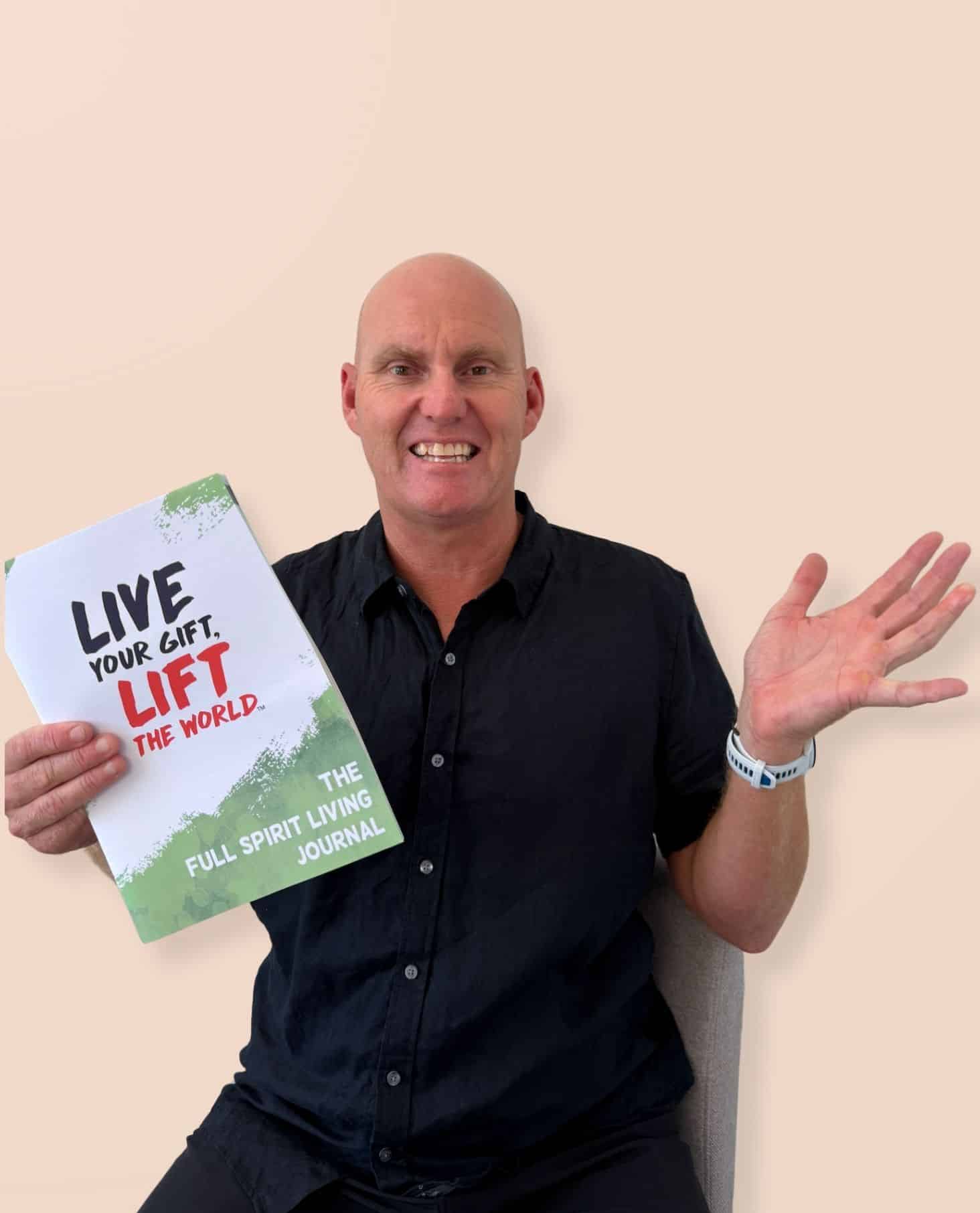 Troy Devine smiling and holding up the Full Spirit Living Journal with the cover reading Live Your Gift, Lift The World
